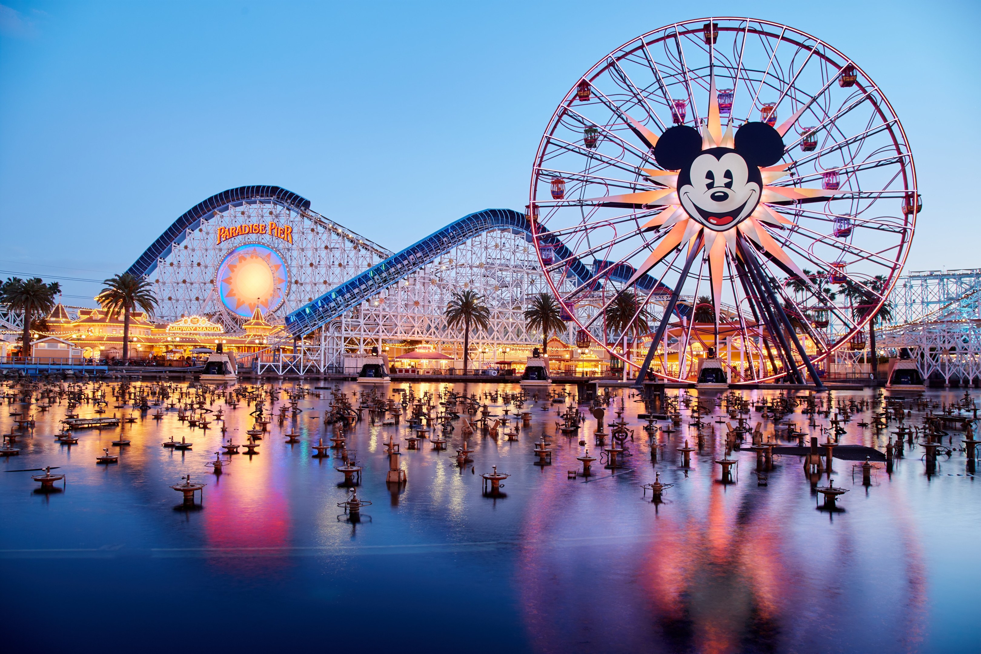 Eine lebendige Dämmerungsszene im Disneyland California Adventure mit einem riesigen Mickey-Maus-Rad im Vordergrund, einer Achterbahn im Hintergrund und Booten auf dem Wasser, mit einem bunten Himmel und silhouettenartigen Bäumen.