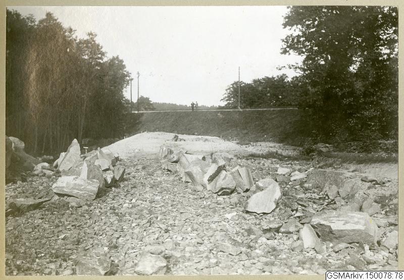 Schwarz-weiß-Foto einer Schotterstraße mit verstreuten Steinen, Bäumen, Strommasten, einer Wand im Hintergrund und Himmel darüber, mit Text, der Routen wie Germantown, New Jersey, New York und New England am unteren Rand auflistet.