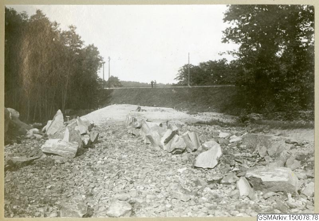 Schwarz-weiß-Foto einer Schotterstraße mit verstreuten Steinen, Bäumen, Strommasten, einer Wand im Hintergrund und Himmel darüber, mit Text, der Routen wie Germantown, New Jersey, New York und New England am unteren Rand auflistet.