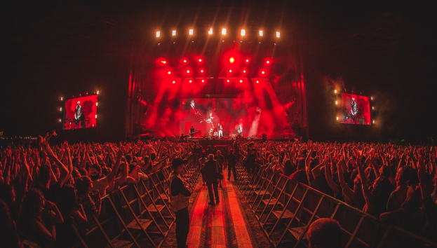 Große Menschenmenge vor einer hell erleuchteten Bühne, auf der Musiker spielen und andere zuschauen, mit dunklem Hintergrund für besseren Kontrast.