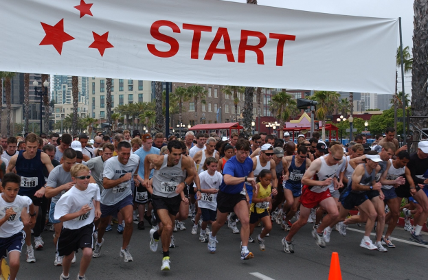 Gruppe von Menschen beim Marathon mit einem Verkehrskegel im Vordergrund und einem Banner mit Text im Hintergrund, umgeben von Bäumen, Laternen, Gebäuden und einem klaren blauen Himmel.