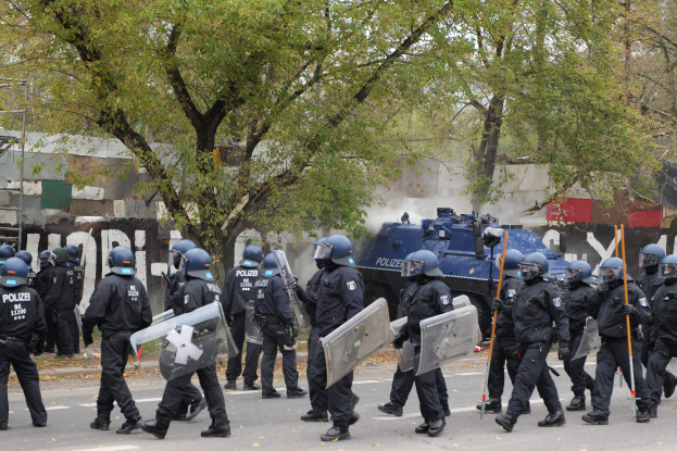 Eine Gruppe von Polizisten in Einsatzausrüstung, die Helme tragen und Schilde und Stöcke halten, geht eine Straße entlang mit einem gepanzerten Fahrzeug, Bäumen, Gebäuden und einem klaren blauen Himmel im Hintergrund.
