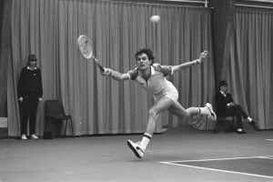 Ein Mann schwingt einen Tennis-Sch├Ąger in Richtung eines Balls, mit vier Personen und Vorh├Ąngen im Hintergrund.