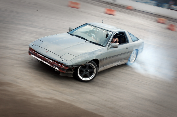 Eine Person mit Helm fährt ein Auto auf einer Strecke, aus dem Rauch aufsteigt, umgeben von orangen Konussen mit einem unscharfen Hintergrund, der hohe Geschwindigkeit andeutet.
