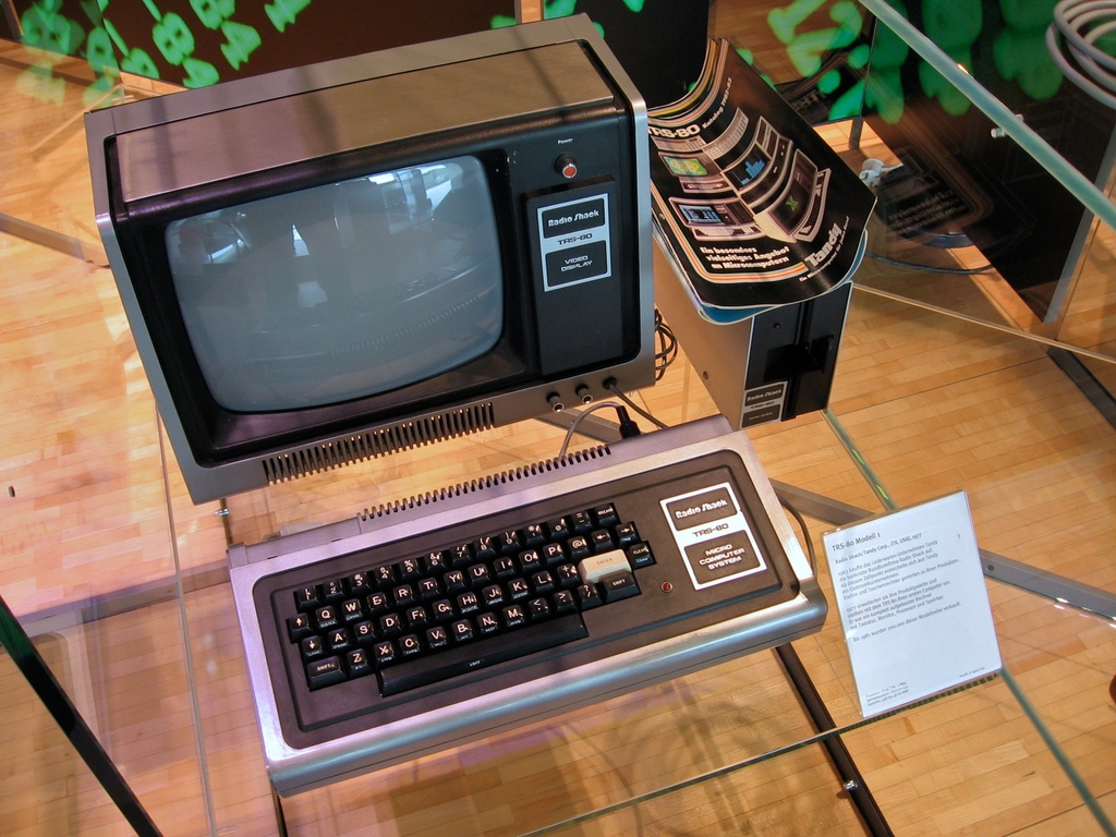 Ein Computersystem auf einem Glastisch mit einer Tafel davor, Broschüren auf der CPU und einem Holzfußboden im Hintergrund.