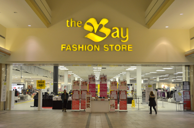 Verkaufsraum eines Bay Fashion Shops in einem Einkaufszentrum mit Kunden, Wandtafeln, Säulen, Deckenleuchten und Hintergrundauslagen.