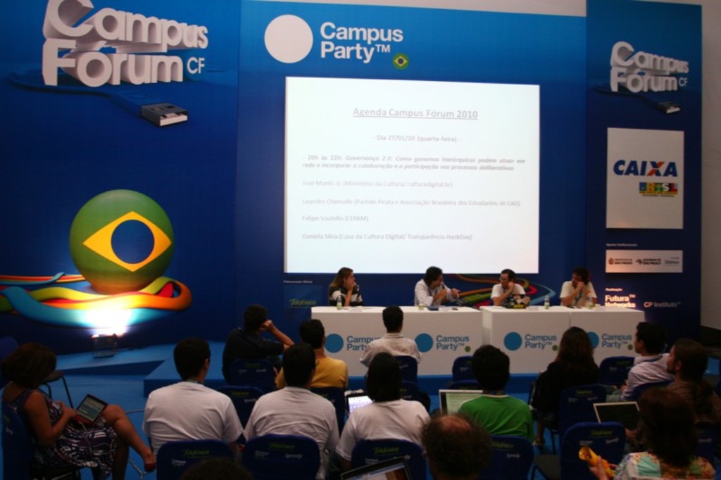 Ein Konferenzsaal mit Teilnehmern an Tischen mit Laptops und Wassergläsern, ein Projektionsschirm mit Inhalt und eine blaue Tafel mit Text auf der rechten Seite.