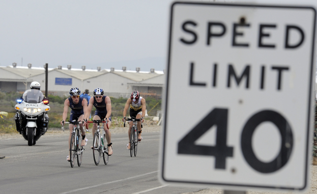 Eine Gruppe von Radfahrern mit Helmen fährt auf einer Straße mit einem Geschwindigkeitsbegrenzungsschild, Gebäuden, Masten, Pflanzen, Bäumen und einem klaren blauen Himmel im Hintergrund.