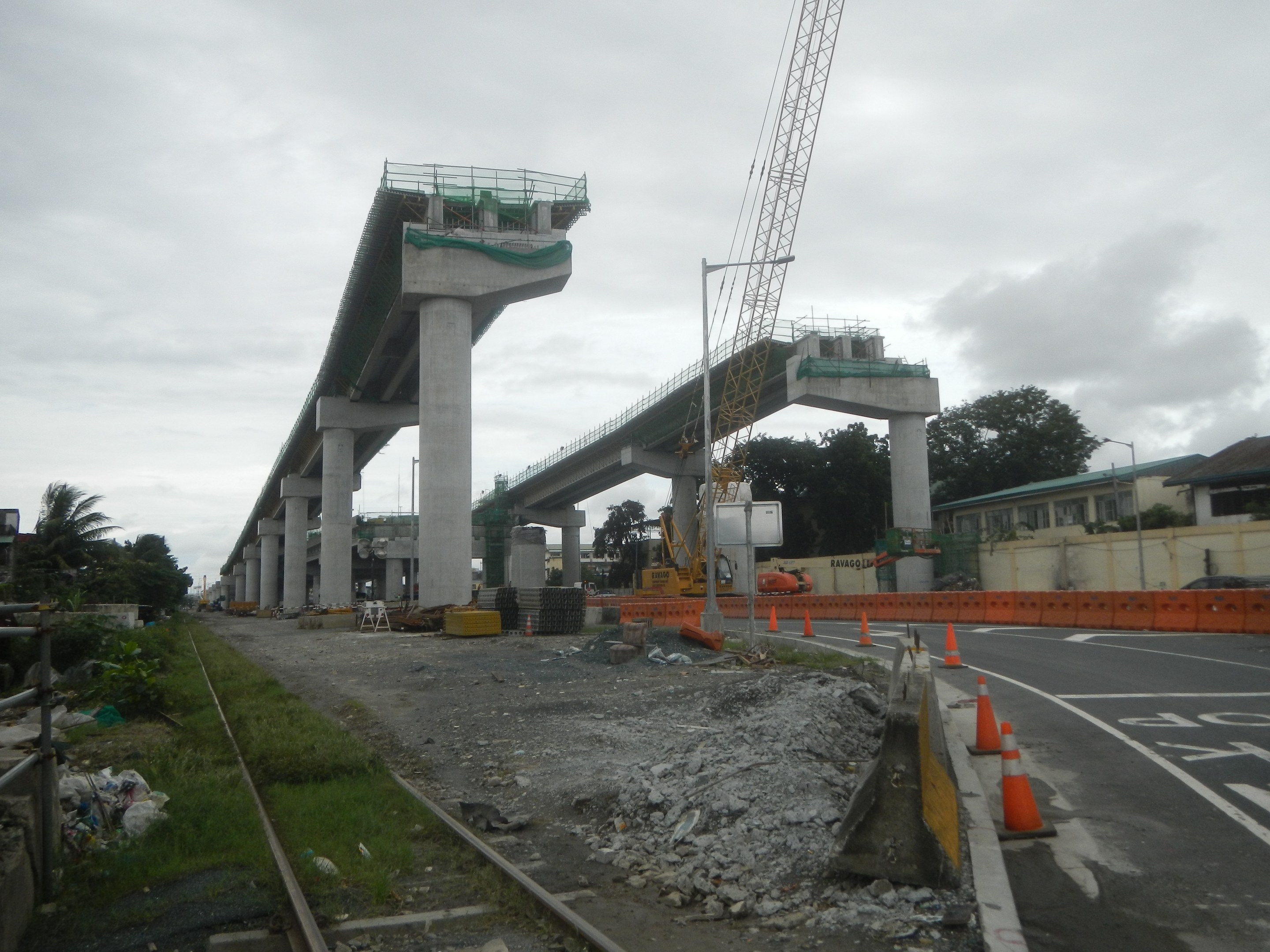 Baustelle mit einer Brücke im Hintergrund, Straße mit Verkehrskegeln markiert, verstreute Steine und Gras, eine Eisenbahnschiene links, Bäume und Gebäude flankieren die Straße und ein bewölkter Himmel darüber.