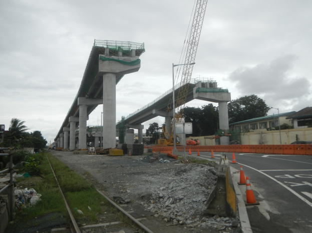 Baustelle mit einer Brücke im Hintergrund, Straße mit Verkehrskegeln markiert, verstreute Steine und Gras, eine Eisenbahnschiene links, Bäume und Gebäude flankieren die Straße und ein bewölkter Himmel darüber.
