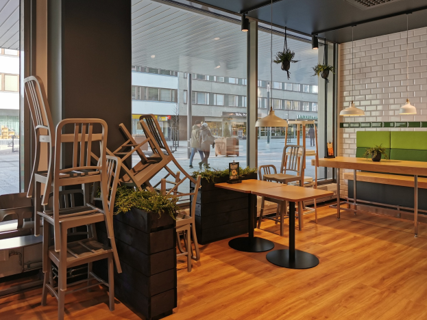 Ein Restaurant-Innenraum mit Holztischen und -stühlen in der Nähe eines großen Fensters, das eine Außenansicht von Berlin mit Passanten, Gebäuden und Straßenelementen zeigt.