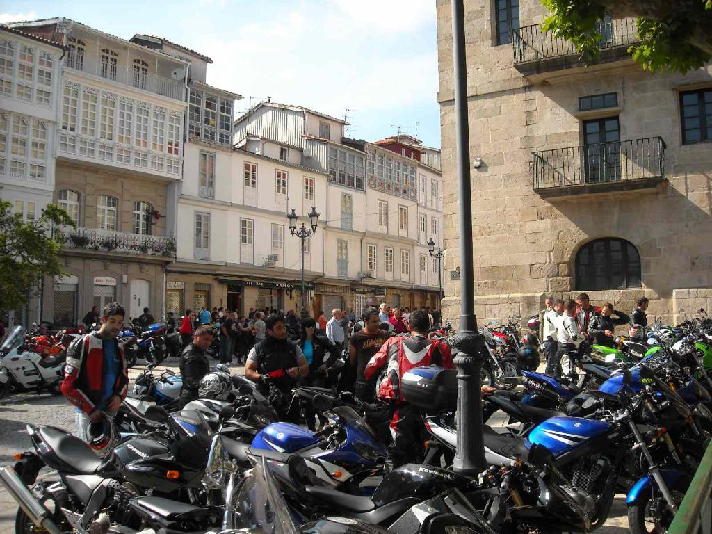 Ein cremefarbenes Gebäude mit vielen Fenstern, das an ein Backsteingebäude mit einer Metallbalustrade angrenzt, mit mehreren geparkten Fahrrädern und Menschen, die davor stehen und sich unterhalten.