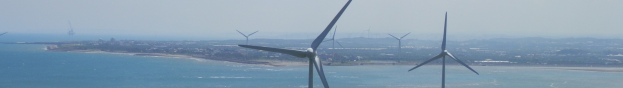 Eine Gruppe von Windrädern im Meer in der Nähe einer Stadt, mit Wasser am Strand, Bäumen und Gebäuden im Hintergrund unter einem klaren blauen Himmel.