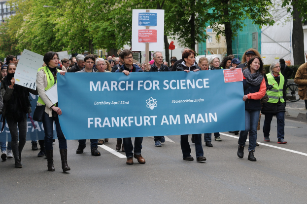 Eine Gruppe von Menschen marschiert auf einer Straße und hält ein "March for Science Frankfurt am Main"-Schild hoch, mit Bäumen und Gebäuden im Hintergrund.