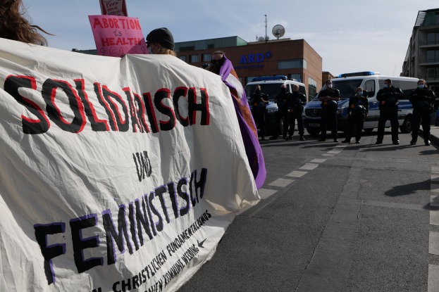 Eine Gruppe von Menschen marschiert auf der Straße und hält ein "Solidarität und Feminismus"-Schild, mit parkenden Fahrzeugen und Gebäuden im Hintergrund unter einem klaren blauen Himmel.