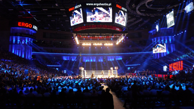 Eine große Menschenmenge in einer Arena schaut in einen Boxring, mit Texttafeln auf beiden Seiten und Bildschirmen und Lichtern an der Decke.