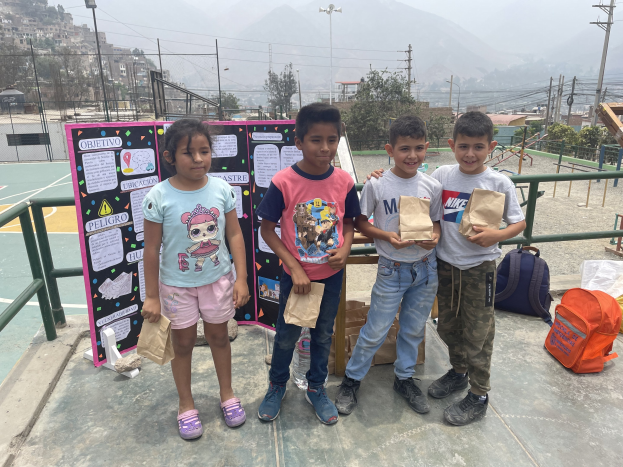 Gruppe von Kindern auf einem Basketballfeld, die Papiersäcke halten, mit Säcken und Gegenständen rechts und einer Tafel mit Text links, vor dem Hintergrund von Zäunen, Masten, Straßenlaternen, Bäumen, Gebäuden, Höhen und Himmel.