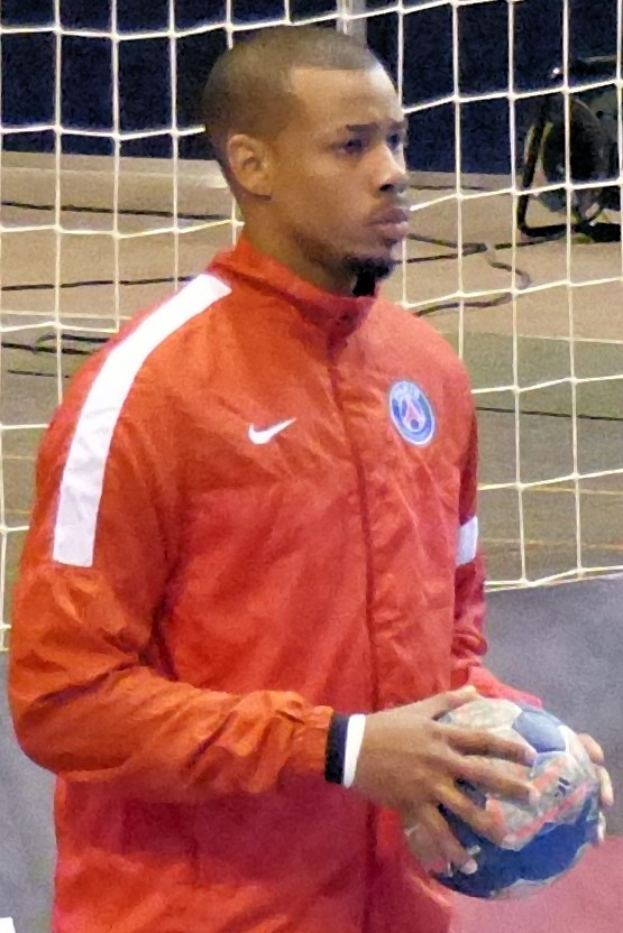 Ein Mann in einem blauen T-Shirt und Shorts steht vor einem Fußballtor und hält entschlossen einen Ball.