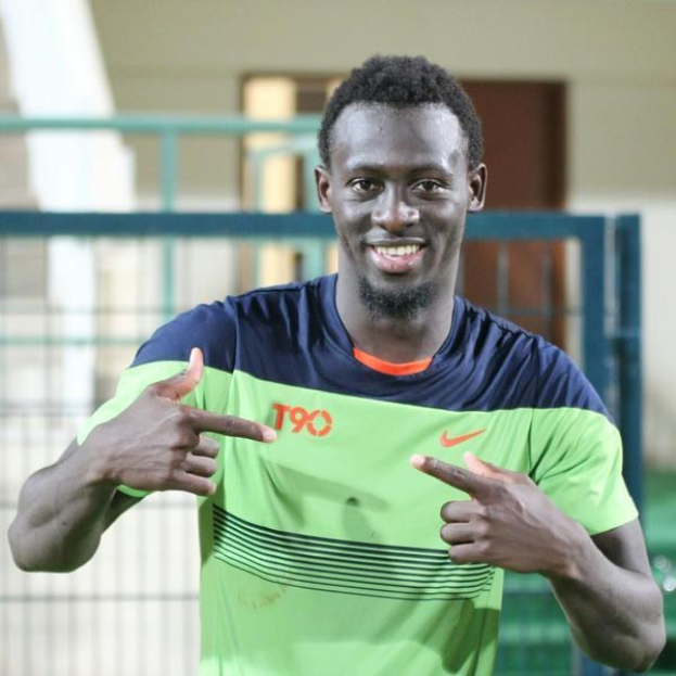 Ein Mann in einem grünen und blauen Hemd zeigt auf die Kamera und lächelt, vor einem unscharfen Geländer, einer Wand und einer Tür.