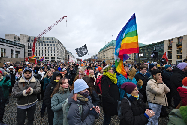 Eine große Gruppe von Menschen bei einer LGBTQ+-Rechtsdemo in Berlin, die Fahnen und Plakate schwenken, mit Gebäuden und einem Kran im Hintergrund unter einem bewölkten Himmel.