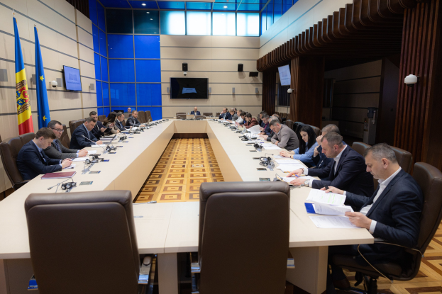 Eine Gruppe von Menschen sitzt um einen langen Tisch mit Papieren, Büchern, Mikrofonen und anderen Gegenständen während einer Sitzung des Europäischen Parlaments in Brüssel. Links sind Flaggen zu sehen, im Hintergrund eine Wand mit Bildschirmen und Lichtern.