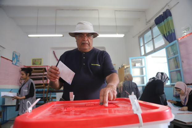 Ein Mann mit Hut stimmt bei einer Wahllokal ab, vor einer roten Wahlurne stehend, während andere sitzen und im Hintergrund schreiben.