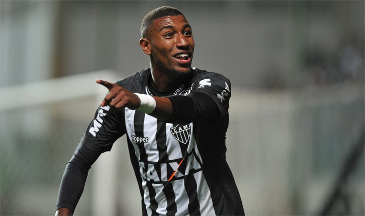 Ein Fußballspieler in einem schwarz-weißen gestreiften Trikot lächelt, während er ein Tor feiert, mit einem unscharfen Hintergrund.