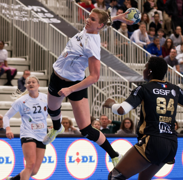 Gruppe von Frauen, die auf einem Platz Handball spielen, mit einem Ball in der Luft, Zuschauern im Hintergrund und einem "Handball-Weltmeisterschaft 2019 - Tag 2"-Schild.