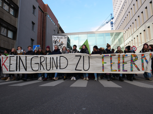 Demonstranten mit einem Banner "Kein Grund zu Feiern" gegen deutsche Sparmaßnahmen, mit Gebäuden, einem Schild, einem weiteren Banner, einem Kran und einem bewölkten Himmel im Hintergrund.