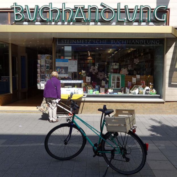 Eine Frau steht neben einem Fahrrad, das vor einem Laden geparkt ist, mit einem Fahrradrad in Vordergrund und durch die Glasscheibe des Ladens sichtbare Bücher.