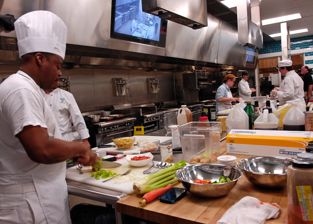 Eine Gruppe von Köchen in einer Küche beim Zubereiten von Speisen auf einem Schneidbrett, mit einem Koch im Vordergrund in einer weißen Schürze und Mütze, der ein Messer hält und von verschiedenen Utensilien, Schüsseln, Gemüse, einem Handy, Flaschen und anderen Gegenständen umgeben ist, mit einem Fernseher und Deckenleuchten im Hintergrund.