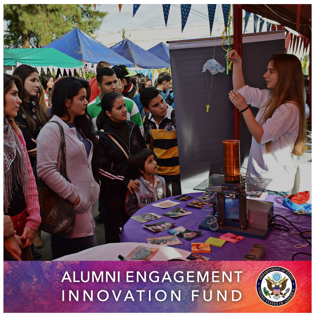 Gruppe von Menschen um einen Tisch versammelt mit einem "Alumni Engagement Innovation Fund"-Schild, umgeben von Gegenständen, mit Zelten und Bäumen im Hintergrund unter einem klaren blauen Himmel.