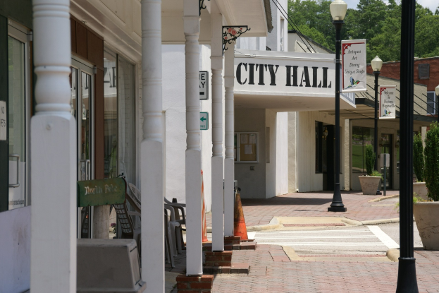 Rathaus mit umliegenden Gebäuden, Straßeninfrastruktur einschließlich Masten, Laternen und Namensschilder, Kraftfahrzeuge auf der Straße, Absperrgitter, verstreute Stühle, Topfpflanzen, Bäume und eine bewölkte Himmel.