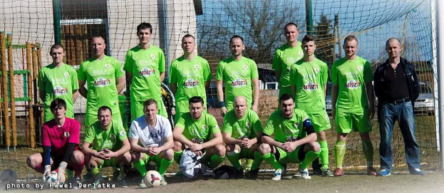 Gruppe von Männern in grünen T-Shirts auf einem Fußballfeld mit Ball und Tor, Bäume und Gebäude im Hintergrund.
