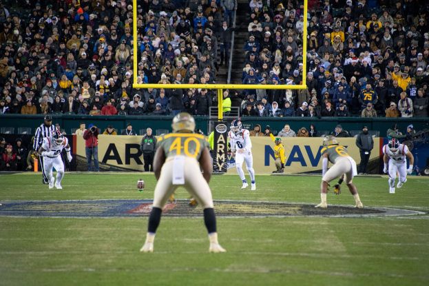 Fußballspieler in Sportkleidung und Helmen konkurrieren auf einem Feld mit Zuschauern im Hintergrund, Stadionstrukturen und sichtbare Beschilderung.