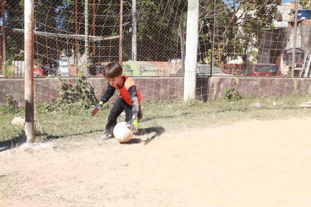 A young boy plays soccer on a dirt field with a cloudy sky, surrounded by grass, plants, poles, a fence, a wall, trees, vehicles, and buildings.