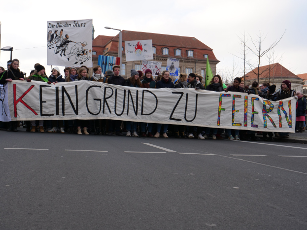 Demonstranten halten ein Transparent mit der Aufschrift "Kein Grund zu Feiern" und stehen auf einer Straße mit Laternenmasten, Straßenlaternen, Bäumen, Gebäuden und einem klaren Himmel im Hintergrund.