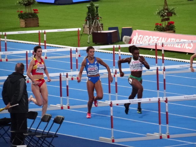 Gruppe von Frauen, die Hürden auf einem Rasenlaufbahn laufen, mit einer Person auf der linken Seite, die einen Stab hält, Stühle und ein Schreibtisch im Vordergrund, Blumentöpfe und Texttafeln im Hintergrund.