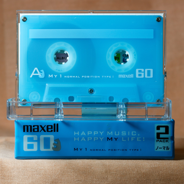Blaue Kassette mit 'Maxell Happy Music, Happy My Life' darauf, auf einer braunen Oberfläche platziert.