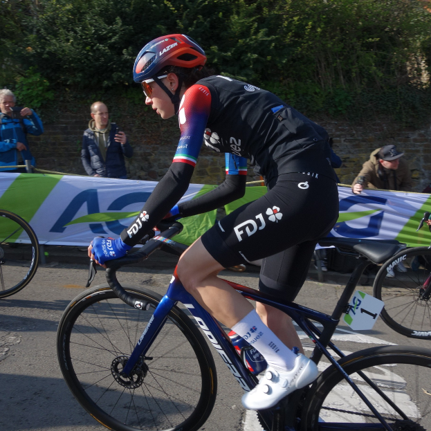 Professionelle Radrennfahrerin Katie Archibald fährt ein Fahrrad auf einer Straße, trägt einen Helm, eine Brille, Handschuhe und Schuhe und fährt an einer Menschenmenge vorbei.