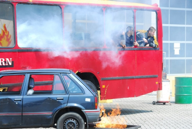 Ein roter Doppeldeckerbus mit Rauch, daneben ein geparktes Auto, drei sichtbare Passagiere im Inneren und ein Gebäude mit Glasfenstern und einem Fass im Hintergrund.
